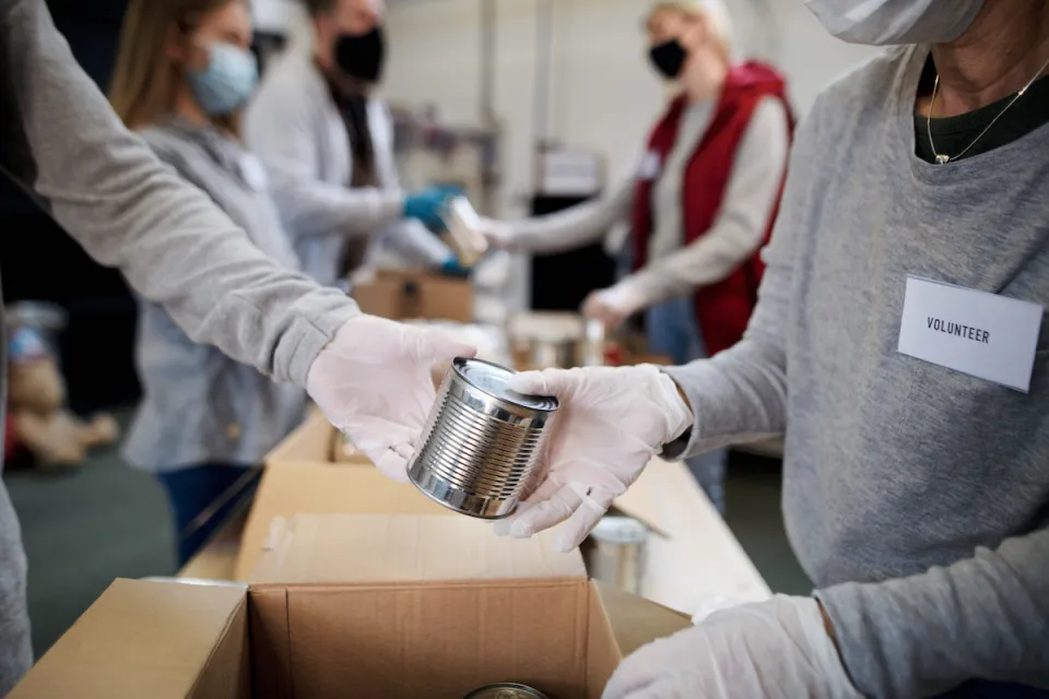 Close-up of people wearing white gloves and Covid-19 masks exchanging unlabeled can of food at food bank